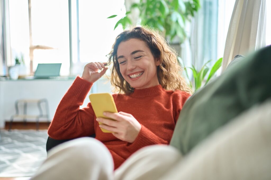 Happy,relaxed young woman sitting on couch using cell phone to shop for christmas phone deals