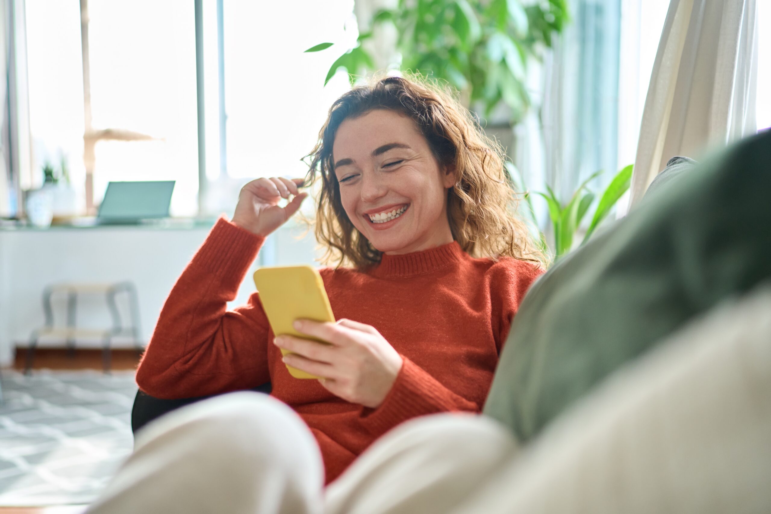 Happy,relaxed young woman sitting on couch using cell phone to shop for christmas phone deals
