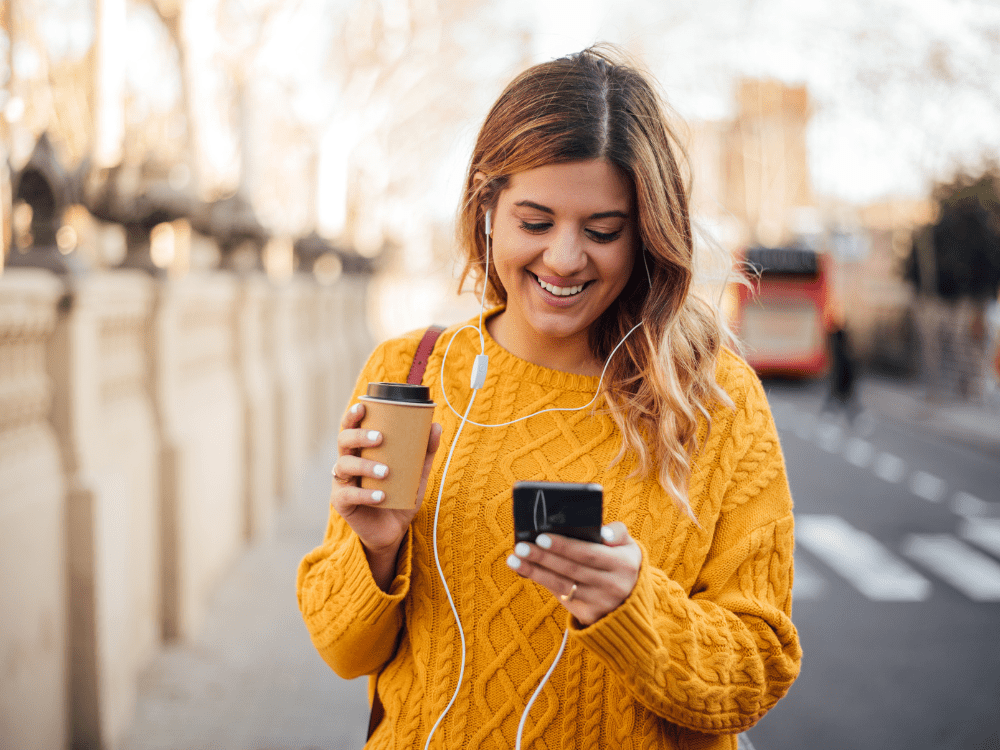The Big Phone Store Honesty Policy Happy woman walking through london with refurbished phone, coffee and earphones